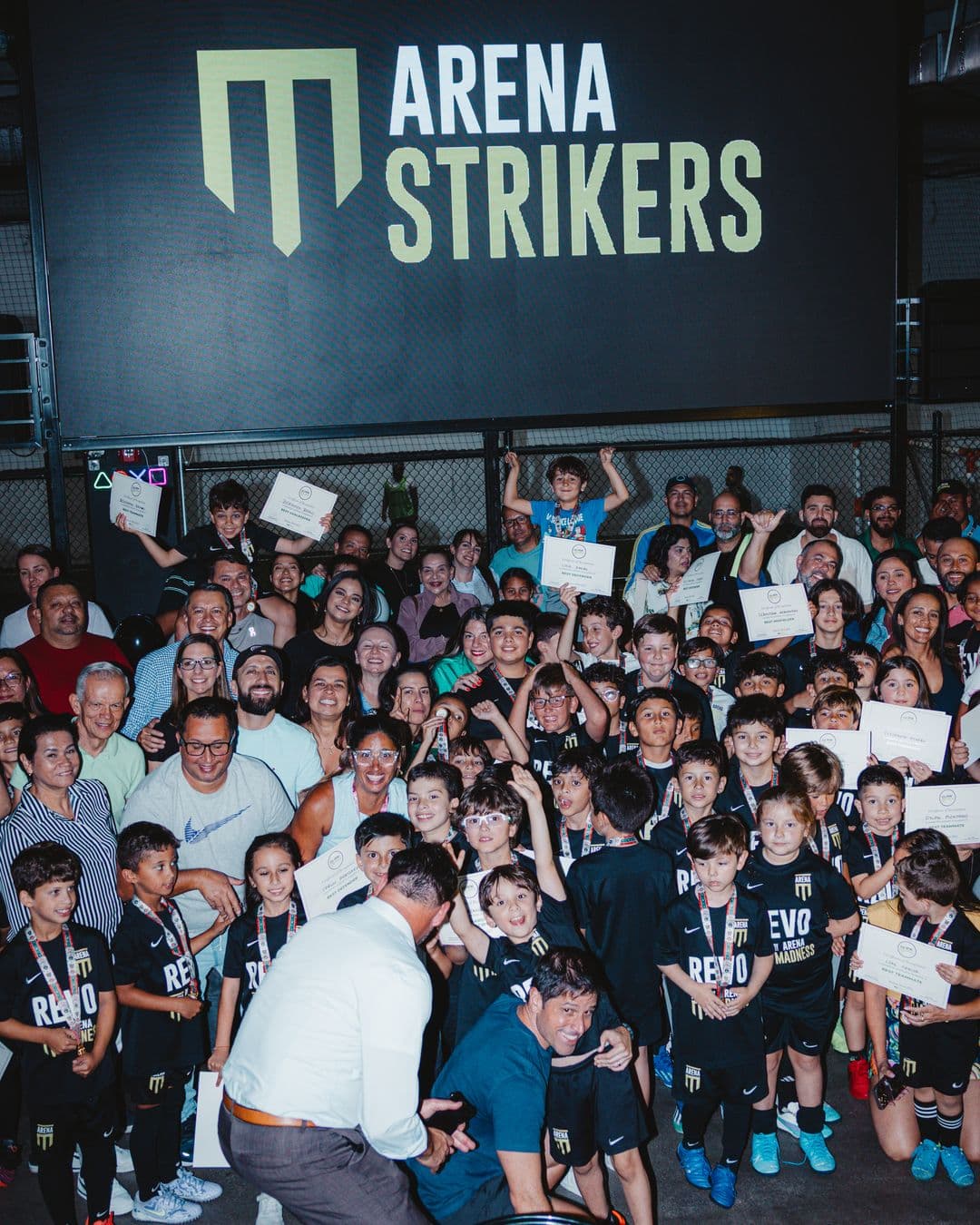 Arena Strikers players holding certificates, with their parents and coaches around them, under the Arena Strikers logo on awards night