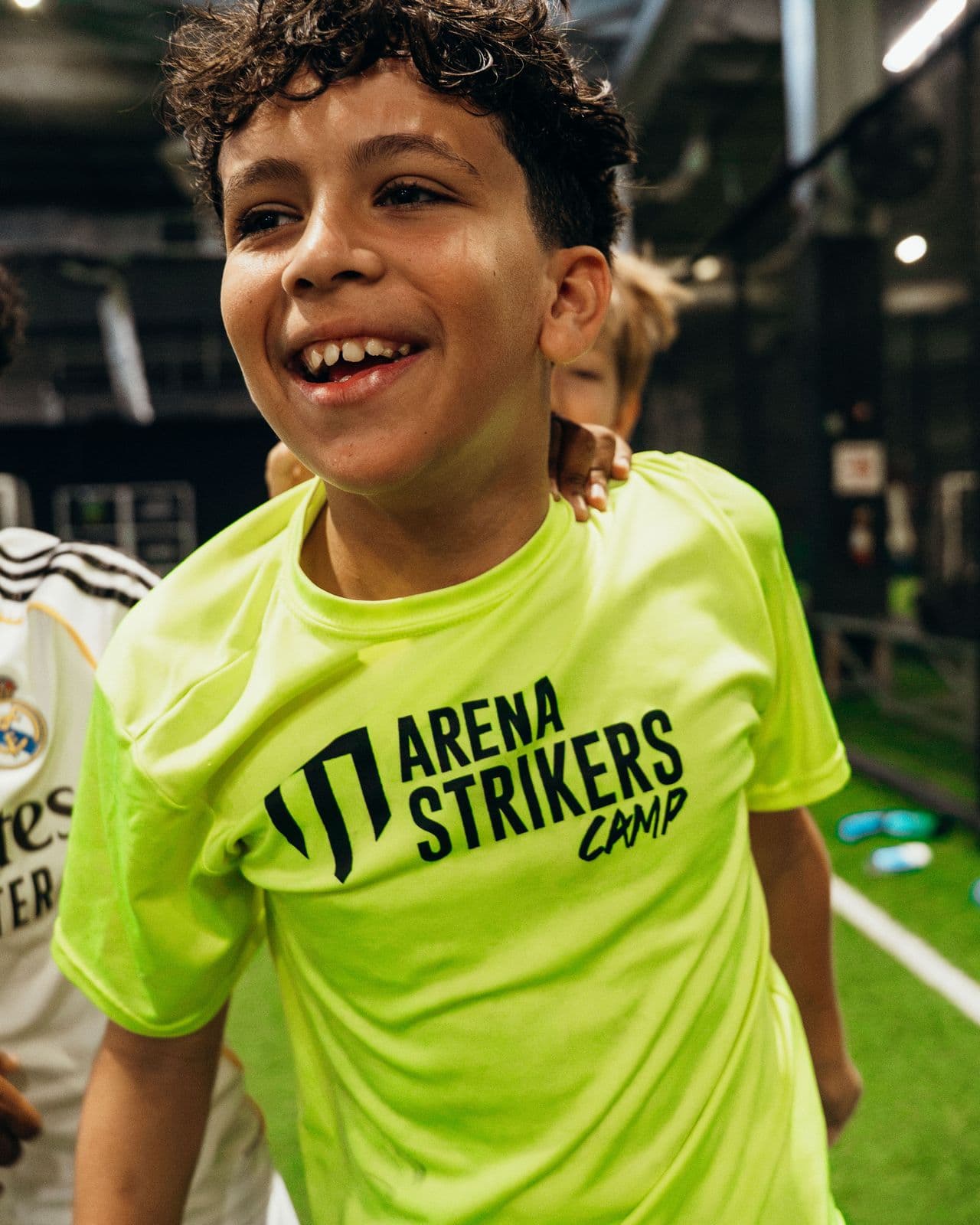 A camper laughing in a lime Arena Strikers Camp shirt on an indoor field