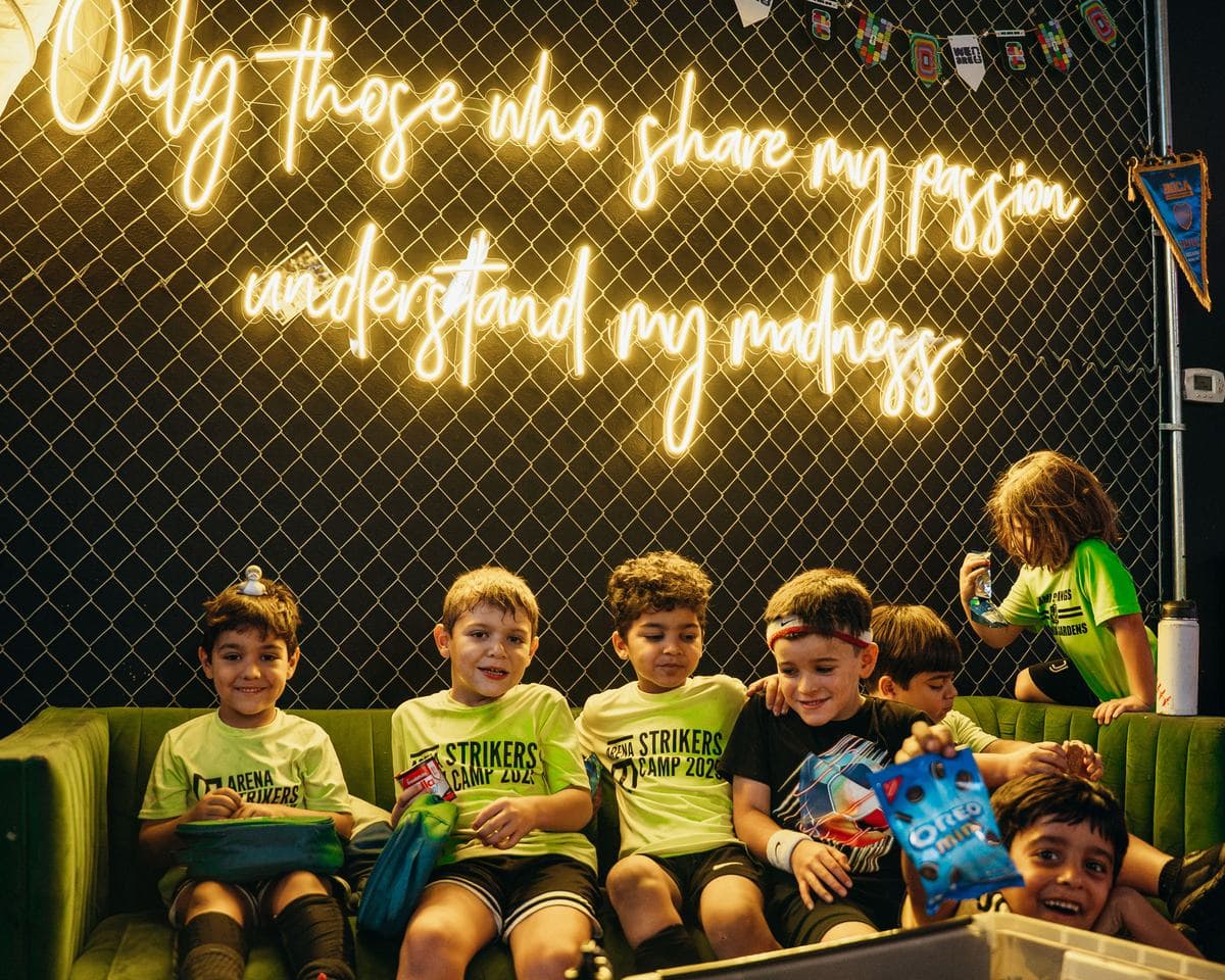 Campers in lime Arena Strikers camp shirts on a green couch under a neon sign that reads "Only those who share my passion understand my madness", one of them holding up a bag of cookies