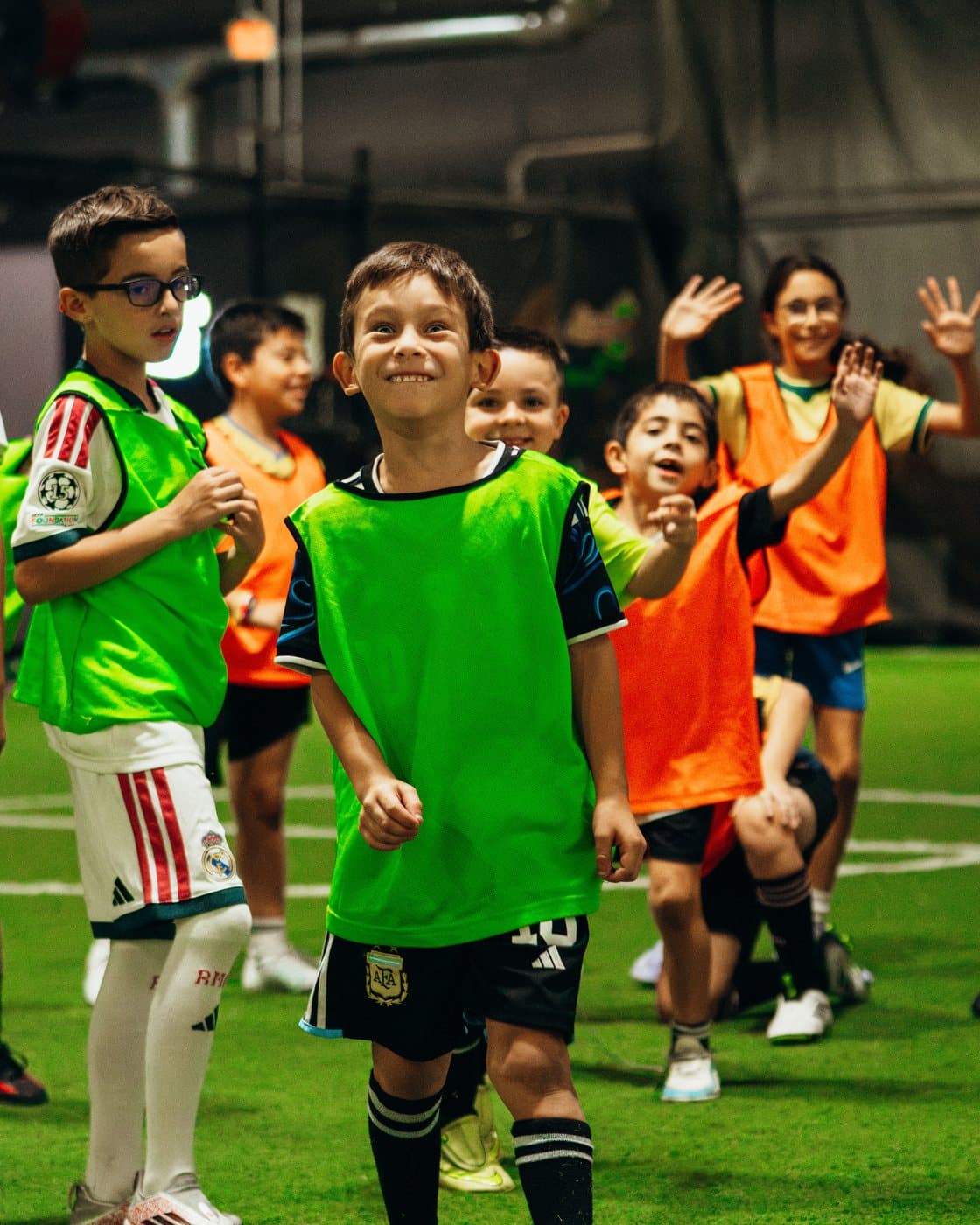 Smiling campers in green and orange bibs on the indoor field, a girl behind them waving with both hands