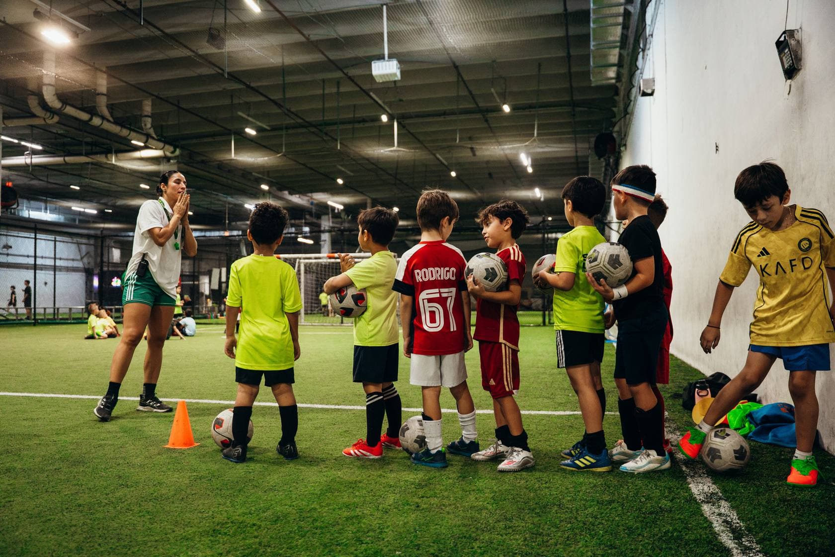 A coach talking to a line of young campers holding soccer balls on an indoor turf field at Arena Strikers' Revo location, summer camp 2026