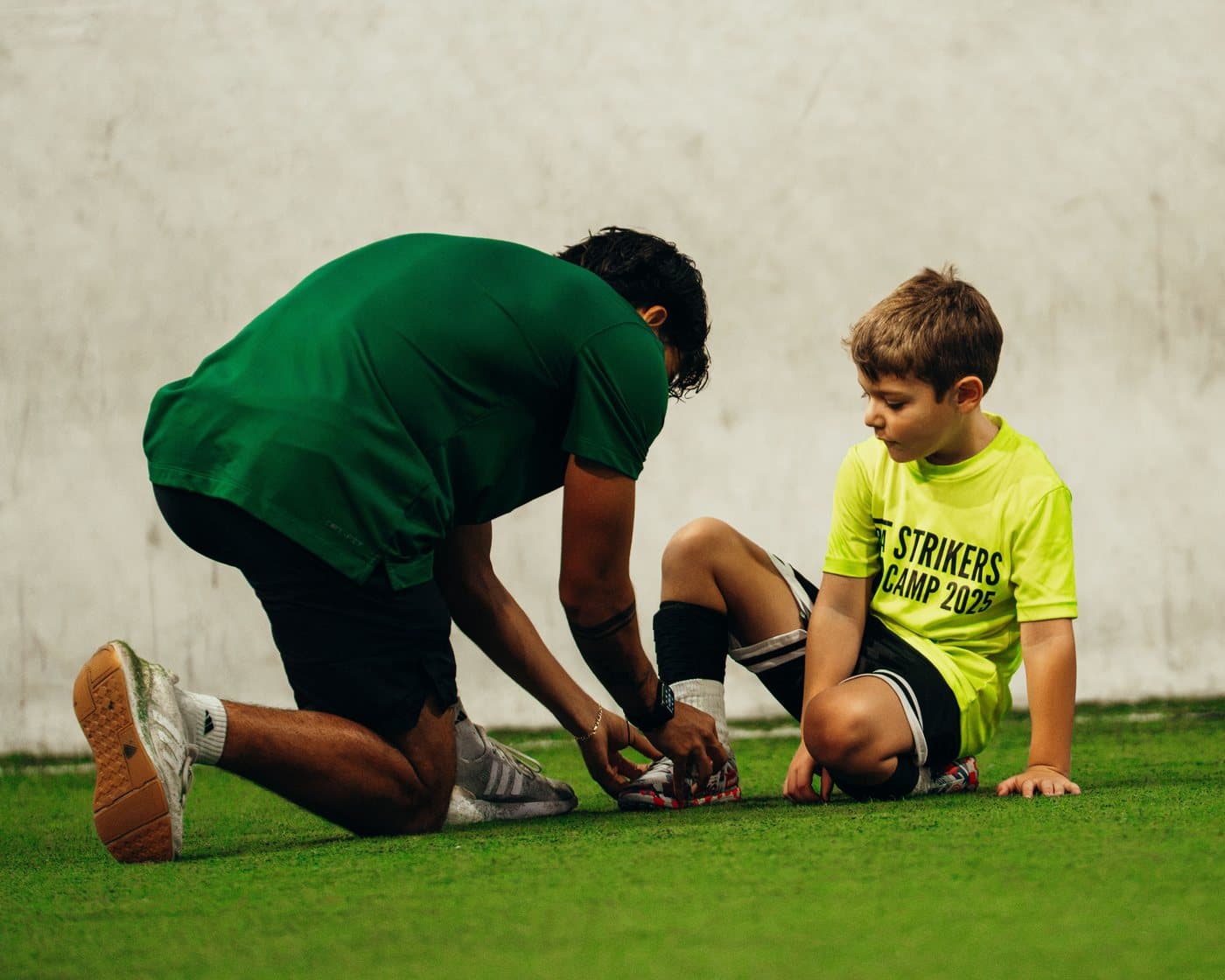A coach kneeling on the indoor turf to tie a camper's shoe