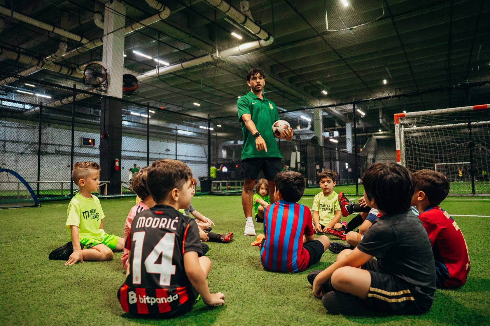 A coach holding a ball talks to young campers sitting around him on the indoor turf at Arena Strikers' Revo location, summer camp 2026