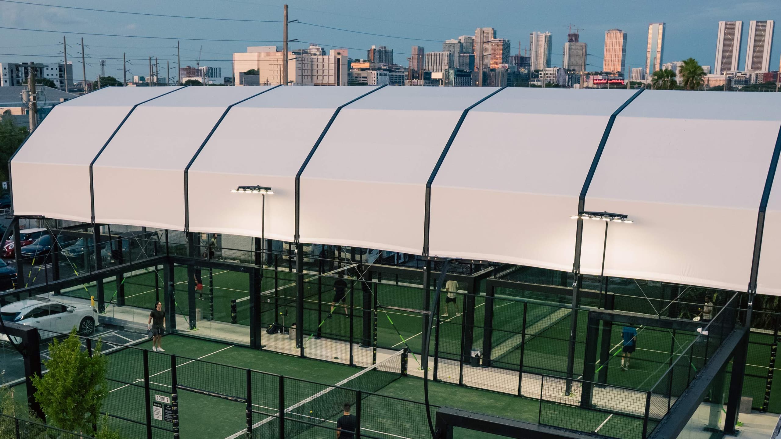 The covered padel courts at Arena SSC Wynwood under their white roof at dusk, with players on court and the Miami skyline behind