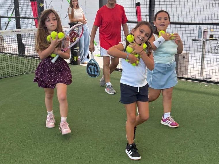 Three girls carrying armfuls of padel balls across a covered court during Kids Academy at Arena SSC Wynwood, Miami