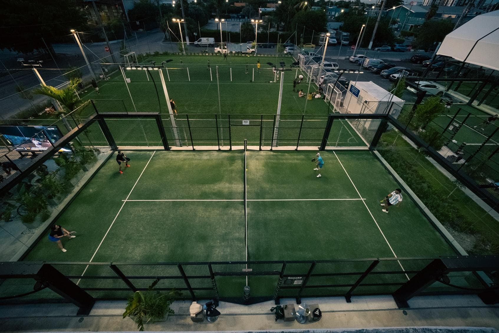 Open-air padel court in play at dusk, seen from above, with the lit soccer field behind, at Arena SSC Wynwood, Miami