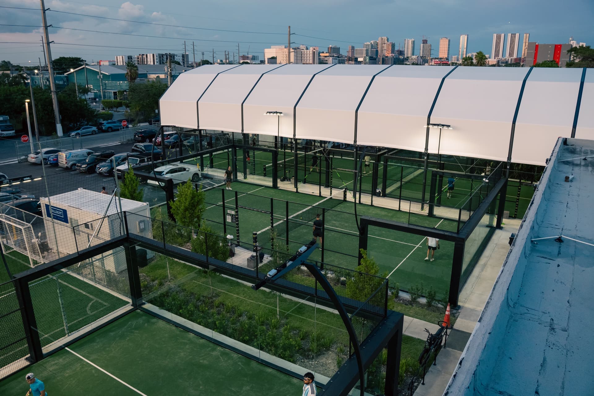 Arena SSC Wynwood from above at dusk: covered padel courts under a white roof, open-air courts and a soccer field, with the Miami skyline behind
